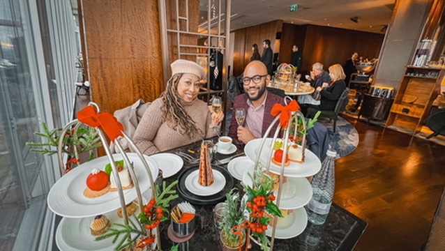 Eulanda and Omo smile at the camera as they hold glasses of champagne behind a table filled with afternoon tea items at TING restaurant, and Shangri-La Hotel London.